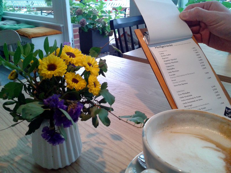 Kaffee, Blumen und Speisekarte im Vordergrund, im Hintergund eine weiße Holzbank